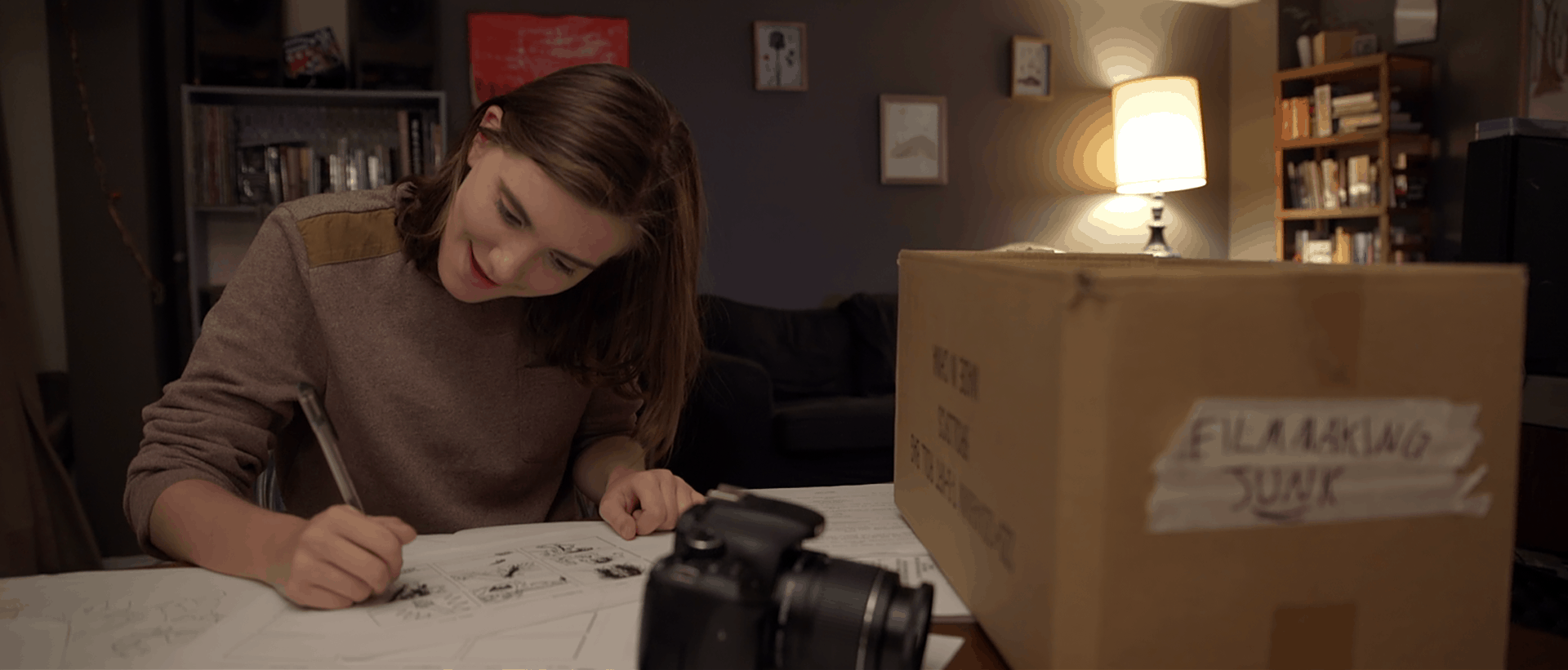 Interview Haley Walker, Actor, Impossible Horror - Haley sits at a table working on storyboards as her character Lily in Impossible Horror. A box sits to her right labelled "Film Stuff". A DSLR camera sits on the table in front of her.
