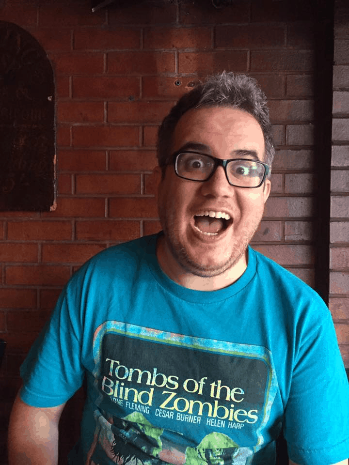 Interview with Justin Decloux, Writer/Director of Impossible Horror - Justin sits in front of a red brick wall and makes a very animated and happy face. He is wearing a blue t-shirt that reads "Tombs of the Zombies". Indie Film Inspiration